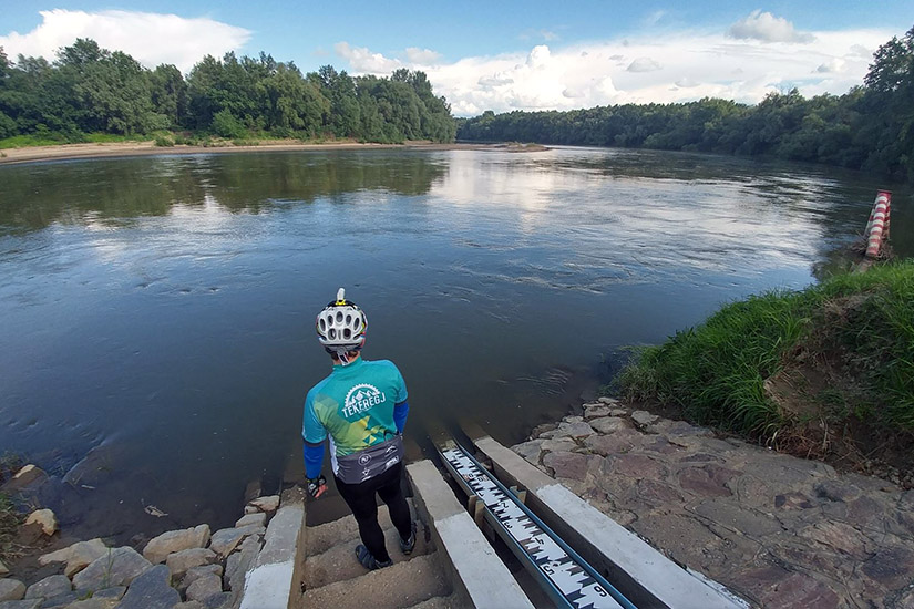 Magyarországi túra: Kerékpáros kék pólóban és sisakban pihen folyóparton, lépcső alján, zöld fák és felhős ég háttérrel. Tekeregj.hu