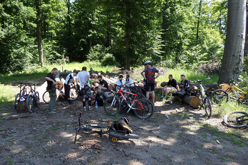 Magyarországi túra: Kerékpárosok pihennek egy erdei tisztáson, a fák között beszélgetnek, kerékpárjaik a közelben, napfény szűrődik át. Tekeregj.hu