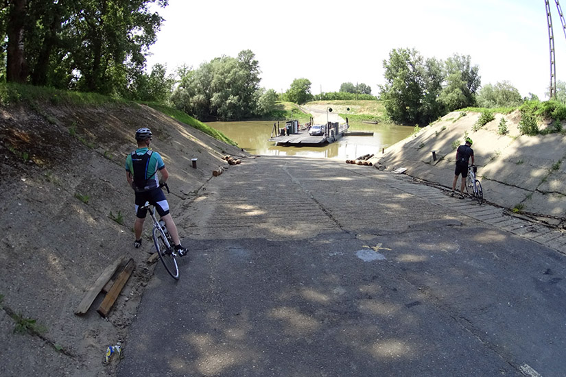 Magyarországi túra: Két kerékpáros a folyóparti révnél, kis komp, zöld fák, napsütéses hegyi túraútvonal festői panorámával. Tekeregj.hu