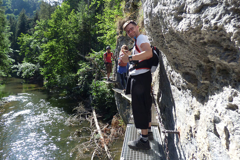 Magyarországi túra: Túrázó mosolyog és mutatja a hüvelykujját egy szurdok feletti vasjárdán, mögötte két ember élvezi a panorámát. Tekeregj.hu