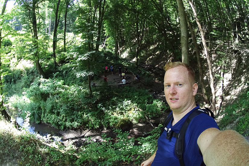 Magyarországi túra: Férfi kék ingben selfie-t készít kiránduláson, napsütötte erdőben, patakparton pihenő túrázókkal a háttérben. Tekeregj.hu