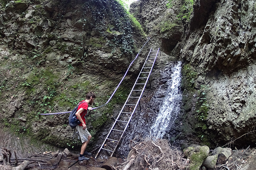 Magyarországi túra: Hátizsákos túrázó fém létra tövében, mohás sziklafal és kis vízesés mellett erdei gyalogtúrán. Tekeregj.hu