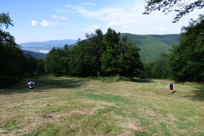 Magyarországi túra: Két túrázó gyalogtúrán egy füves domboldalon, fák és erdős hegyek közt, völgy panorámával, részben felhős ég alatt. Tekeregj.hu