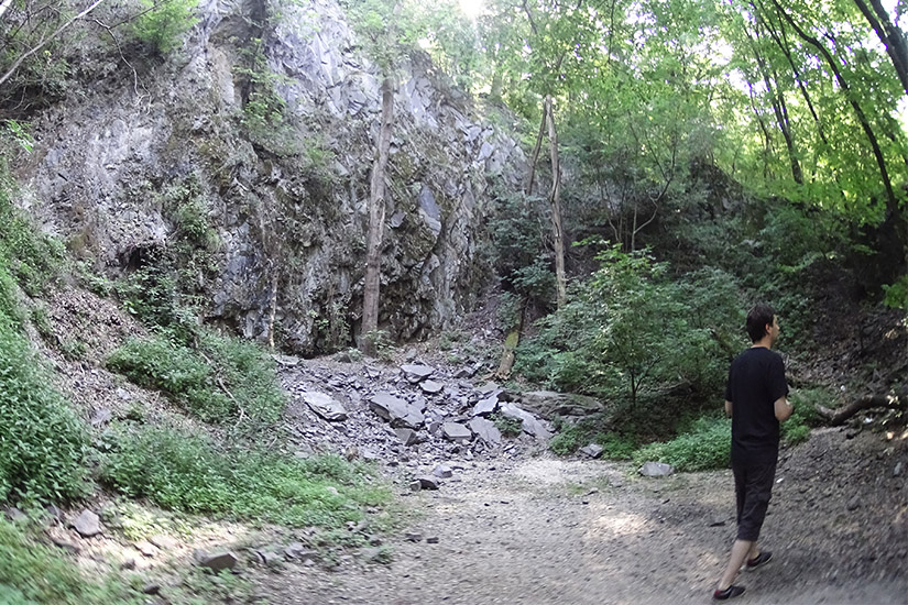 Magyarországi túra: Kiránduló gyalogtúrázik erdei ösvényen, zöld fák, növényzet és sziklás partszakasz mellett. Outdoor élmény Magyarországon. Tekeregj.hu