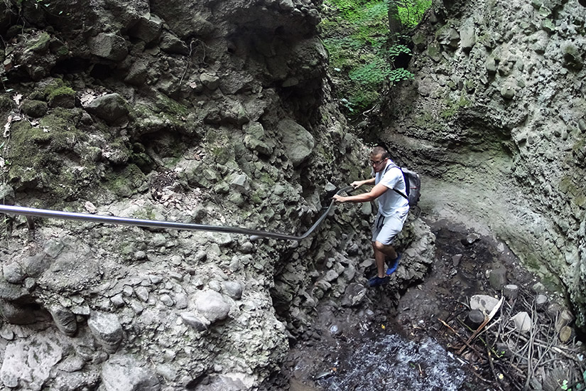 Magyarországi túra: Túrázó hátizsákkal kapaszkodik fel sziklás ösvényen, korlát mellett, mohás sziklafalak és zöld növényzet között. Tekeregj.hu