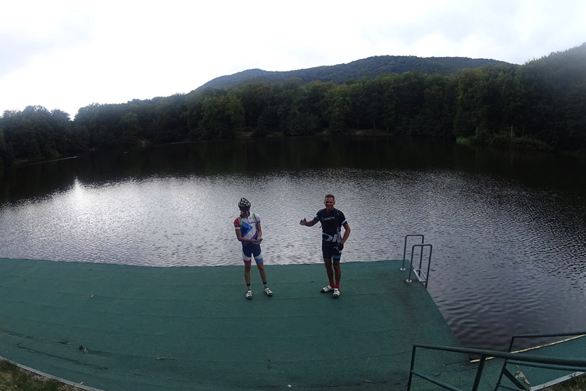 Magyarországi túra: Két biciklis áll egy zöld stégen, nyugodt tóparton, erdős dombokkal a háttérben, felhős ég alatt - természetjárás. Tekeregj.hu