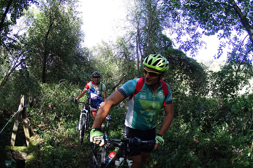 Magyarországi túra: Két biciklis bukósisakban és túraöltözetben teker egy erdei ösvényen, hegyi túra zöld környezetben. Tekeregj.hu