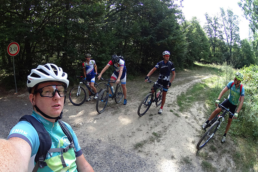 Magyarországi túra: Öt biciklis megáll erdei hegyi túra közben, egyikük szelfit készít, a többiek pihennek a fák és útjelző tábla mellett. Tekeregj.hu