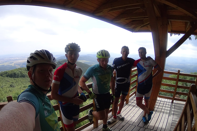Magyarországi túra: Öt kerékpáros sportfelszerelésben pózol a hegyi túra erkélyén, panorámás kilátással a dombokra és mezőkre. Tekeregj.hu
