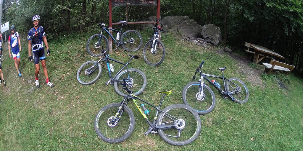 Magyarországi túra: Öt mountain bike fekszik füvön, három túrázó áll mellette; erdő, piknikasztal és hegyi kirándulás tábla a háttérben. Tekeregj.hu