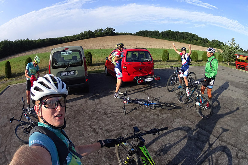 Magyarországi túra: Színes nyugás kerékpárosok készülődnek hegyi túrára parkolóban, egyikük szelfit készít, háttérben napos panoráma. Tekeregj.hu