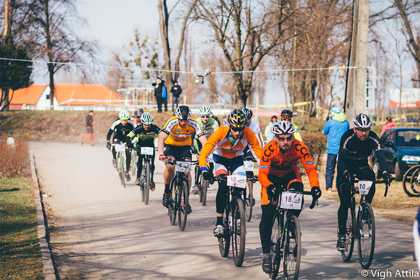 Magyarországi túra: Kerékpáros versenyzők sisakban és rajtszámmal haladnak aszfaltos ösvényen, nézők figyelik a természetben. Tekeregj.hu
