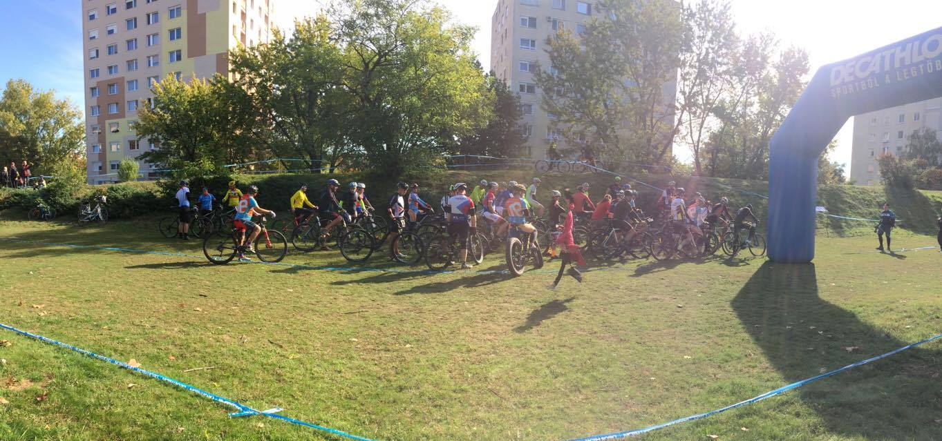 Magyarországi túra: Kerékpárosok sorakoznak a rajtnál füves téren, fák és magas házak között, kék felfújható kapu alatt, napsütésben. Tekeregj.hu