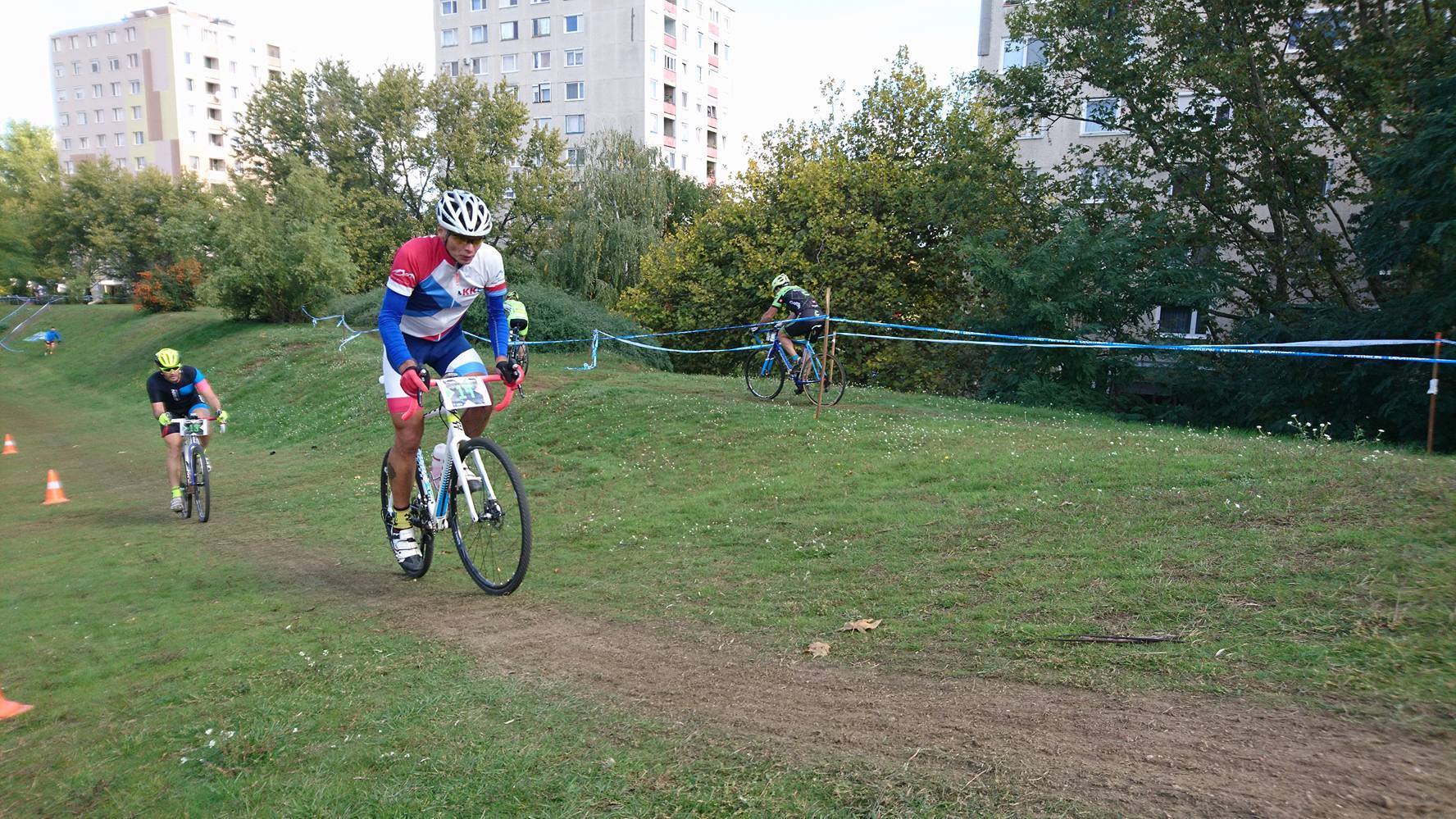Magyarországi túra: Színes nyugás kerékpárosok versenyeznek füves ösvényen, kis emelkedőn, városi panorámával háttérben, jelzőszalagokkal. Tekeregj.hu