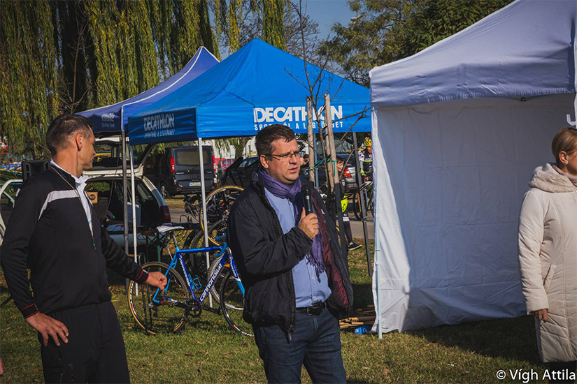 Magyarországi túra: Férfi mikrofont tartva beszél sátrak és kerékpárok mellett egy kiránduláson, napos, fás környezetben. Tekeregj.hu