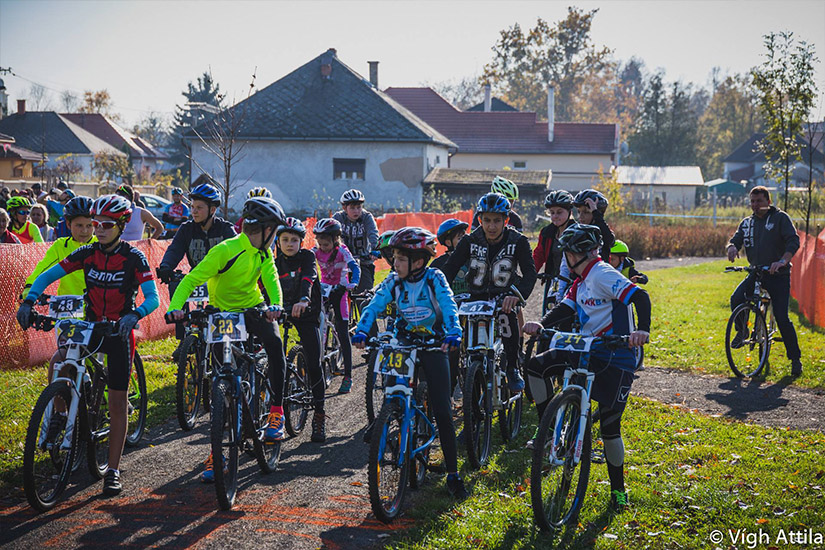 Magyarországi túra: Fiatal kerékpárosok rajtnál sisakkal, mezben, napsütéses versenyen falusi panorámával; hozás esemény. Tekeregj.hu