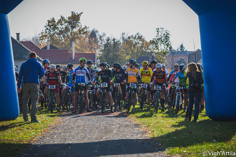 Magyarországi túra: Kerékpárosok színes mezben, sisakban várják a túra rajtját kék felfújható kapu alatt, nézők fotóznak a helyszínen. Tekeregj.hu
