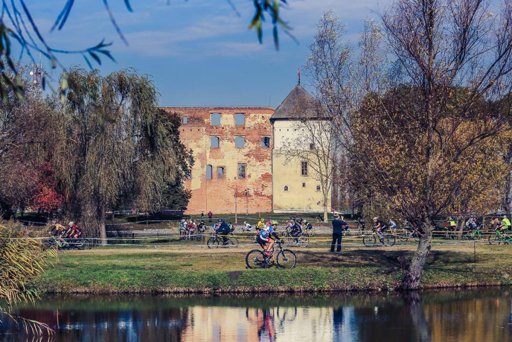 Magyarországi túra: Kerékpárosok túráznak egy tóparti ösvényen, háttérben romos téglaépület és lombtalan fák alatt kék égbolt. Tekeregj.hu