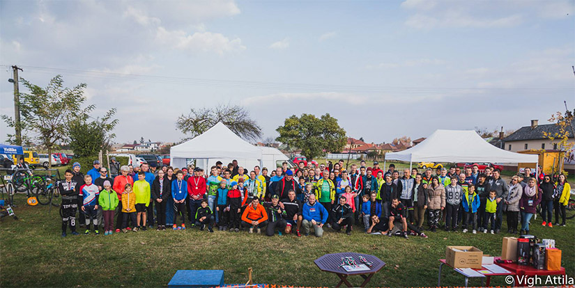 Magyarországi túra: Kiránduló csoport színes sportruhában, gyerekek és felnőttek sátrak előtt, felhős ég alatt közös fotón a természetben. Tekeregj.hu