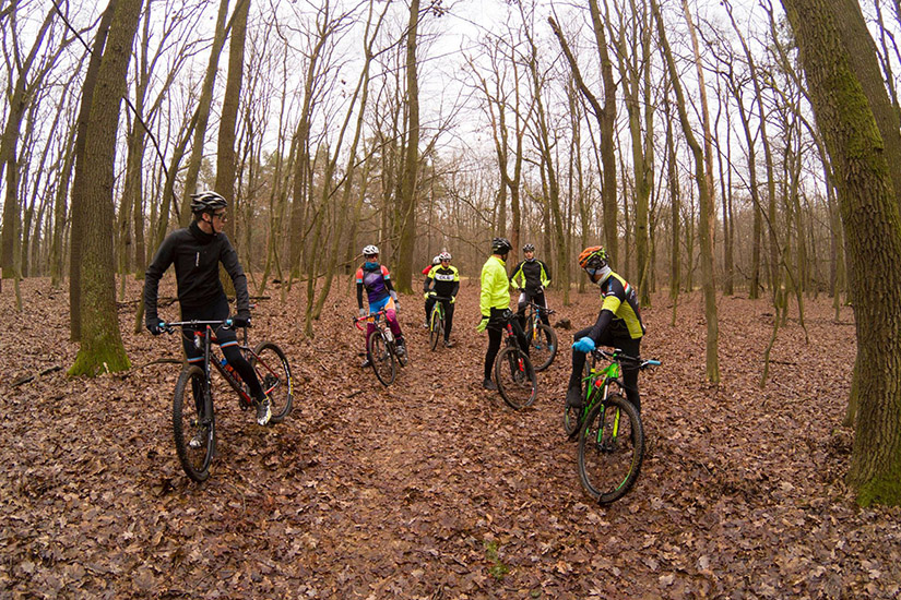 Magyarországi túra: Hegyi kerékpározók színes felszerelésben pihennek egy lombtalan, erdei túraútvonalon borús időben. Tekeregj.hu