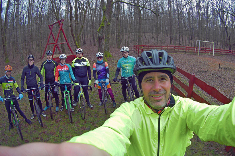 Magyarországi túra: Kerékpáros csoport sisakban és színes ruhában egy erdei tisztáson, selfie-t készítenek, körülöttük fák, fa kerítés. Tekeregj.hu