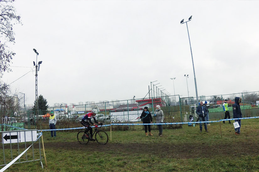 Magyarországi túra: Kerékpáros piros mezben és sisakban teker sáros mezőn egy versenyen, nézők a kordonnál, felhős időben. Tekeregj.hu