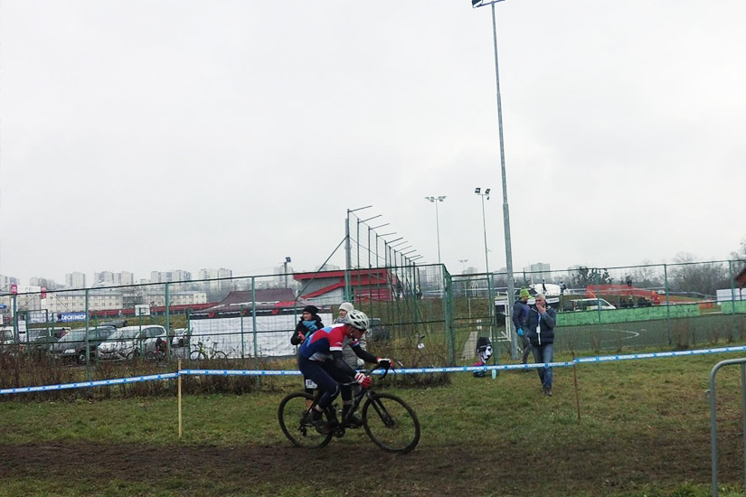 Magyarországi túra: Kerékpáros vörös-fehér mezben teker sáros ösvényen versenyen, nézők és autók a háttérben borús időben. Tekeregj.hu