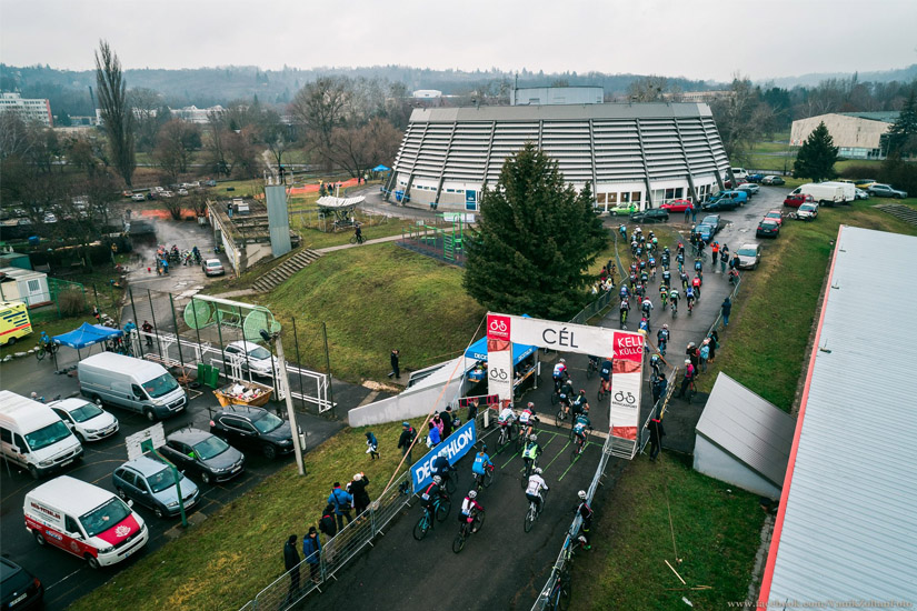 Magyarországi túra: Kerékpárosok érkeznek a CÉL feliratú kültéri célvonalhoz, nézők és autók mellett, felhős túranapon. Tekeregj.hu