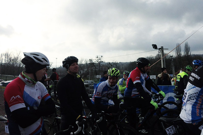 Magyarországi túra: Kerékpárosok színes felszerelésben várakoznak biciklijeikkel egy verseny rajtvonalánál, fák között, felhős időben. Tekeregj.hu