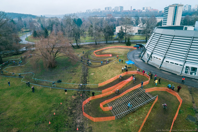 Magyarországi túra: Légifotó kanyargós kerékpárpályáról narancssárga korlátokkal, versenyzőkkel, modern épülettel és zöld környezettel. Tekeregj.hu