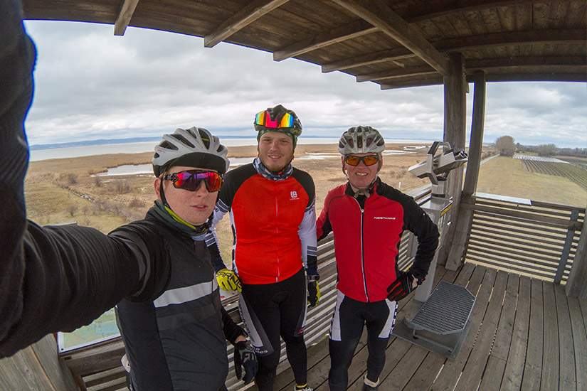 Magyarországi túra: Három biciklis sisakban, napszemüvegben szelfizik egy fa kilátón hegyi túrán, vízparti panorámával a háttérben. Tekeregj.hu