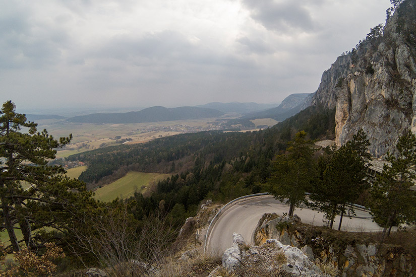 Magyarországi túra: Kanyargós hegyi út sziklafal mentén, fenyőkkel és távoli faluval-ideális gyalogtúrához a természetben. Tekeregj.hu