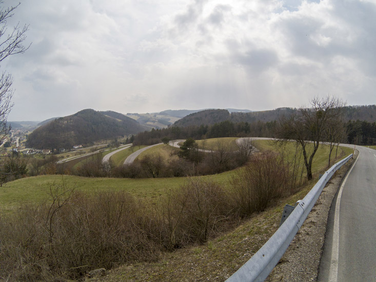 Magyarországi túra: Kanyargós út dombos vidéken, füves és kopár fákkal, felhős ég alatt - ideális gyalogtúrához vagy kerékpározáshoz. Tekeregj.hu