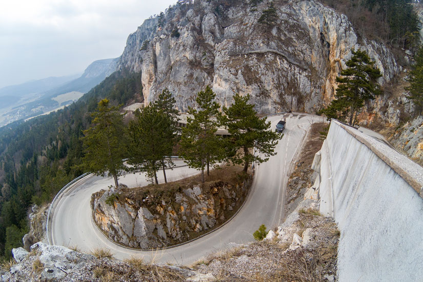 Magyarországi túra: Kanyarodó hegyi út sziklás falak közt, fenyőfákkal, korláttal; túraútvonal felhős ég alatt, meredek panorámával. Tekeregj.hu