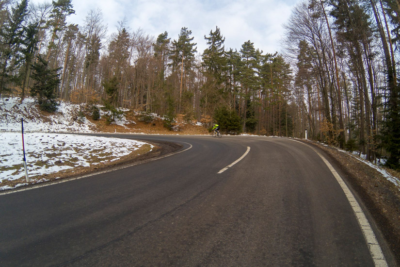 Magyarországi túra: Kerékpáros teker egy kanyargós hegyi úton, hófoltok és magas fák között, felhős időben - szabadtéri kerékpározás. Tekeregj.hu