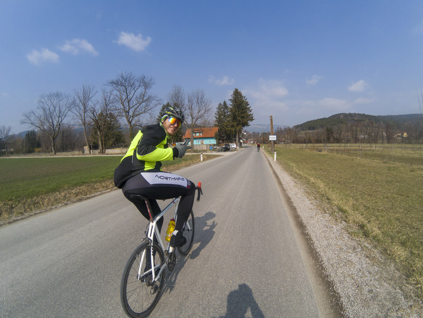 Magyarországi túra: Kerékpáros zöld-fekete szerelésben vidéki úton túrázik, mosolyogva integet, háttérben mezők, dombok és tiszta kék ég. Tekeregj.hu