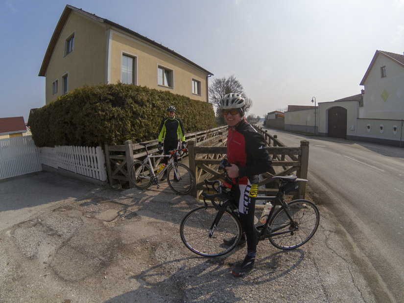 Magyarországi túra: Kerékpárosok túrafelszerelésben egy ház és fakerítés mellett, napsütésben, panorámás kilátással. Tekeregj.hu
