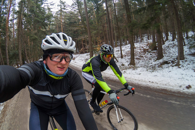Magyarországi túra: Két kerékpáros bukósisakban és téli ruhában havas, erdős úton hegyi túrázik, gyönyörű panoráma a háttérben. Tekeregj.hu
