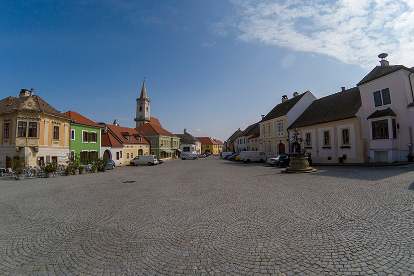 Magyarországi túra: Tágas, macskaköves tér gyalogtúrázáshoz, színes házak, templomtorony és szökőkút alatt részben felhős ég. Tekeregj.hu
