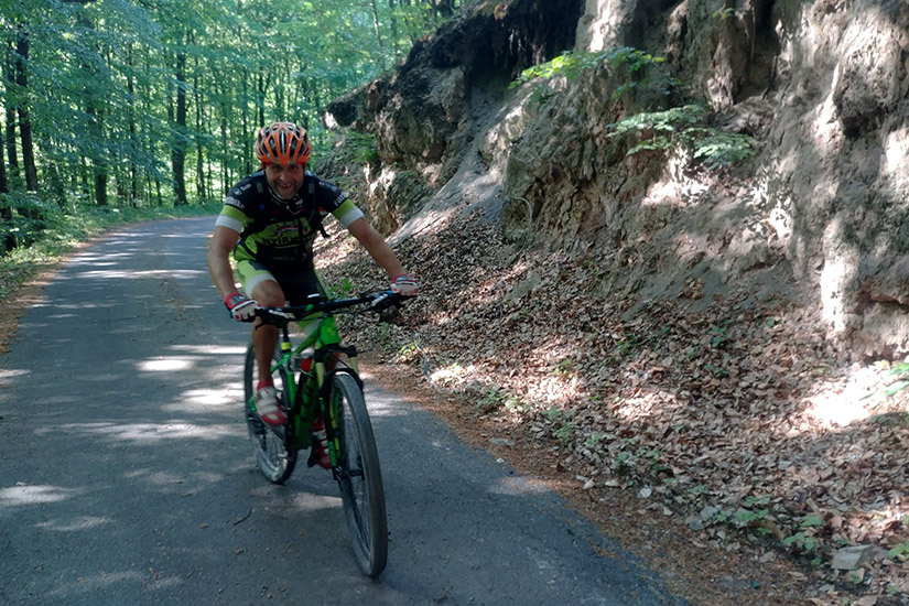 Magyarországi túra: Kerékpáros bukósisakban és sportfelszerelésben teker egy zöld mountain bike-kal erdei aszfaltúton, napsütésben. Tekeregj.hu