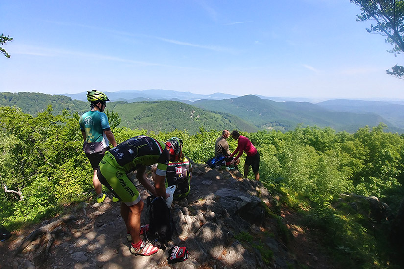 Magyarországi túra: Kerékpárosok felszerelésben egy sziklás kilátón, zöld erdő és hegyek panorámájával a tiszta ég alatt. Tekeregj.hu