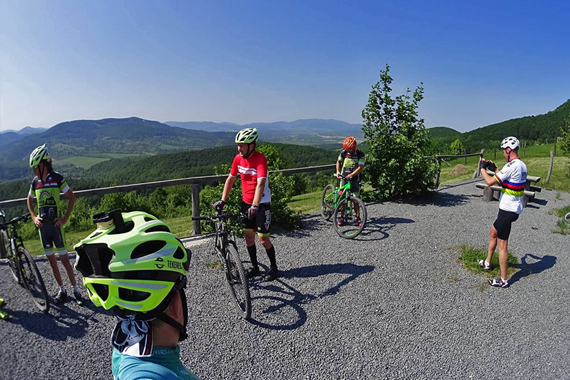 Magyarországi túra: Kerékpárosok hegyi túrán pihennek zöld dombok és hegyek panorámájánál, egyikük fényképet készít. Tekeregj.hu