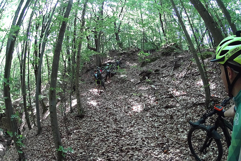 Magyarországi túra: Kerékpárosok tolják biciklijüket egy meredek, erdős ösvényen, napsütésben. Elöl sárga sisakos hozás halad. Tekeregj.hu