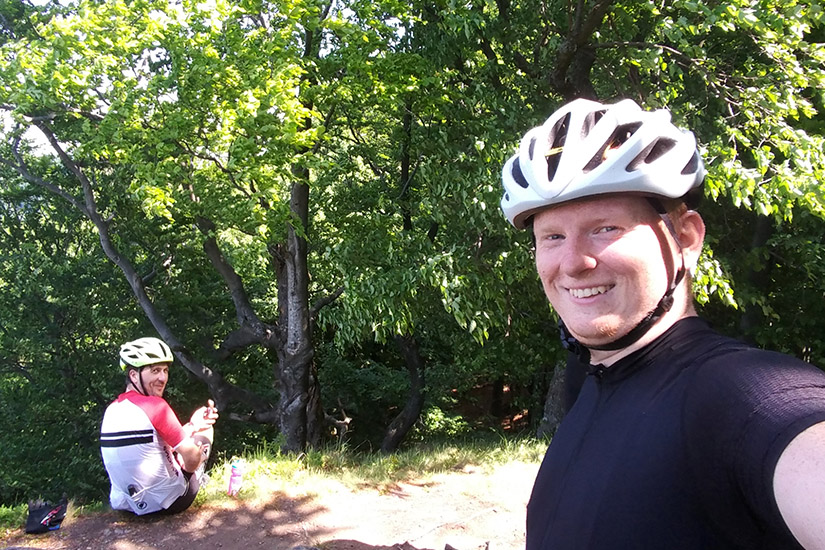 Magyarországi túra: Két bukósisakos kerékpáros pihen napos, erdei kiránduláson, egyikük kamerába mosolyog, másikuk telefonját nézi. Tekeregj.hu