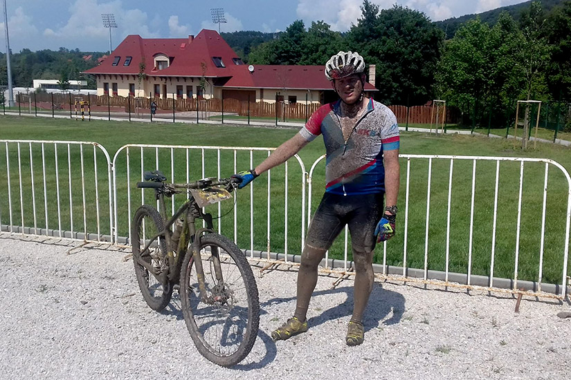 Magyarországi túra: Sáros kerékpáros a hegyi túra után, kavicsos úton, háttérben zöld rét, fehér kerítés és piros tetős épület. Tekeregj.hu