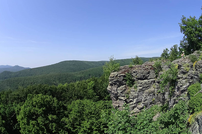 Magyarországi túra: Sziklás szirt sűrű zöld erdőben, tiszta ég alatt, dombok a háttérben - ideális panoráma hegyi túrázáshoz. Tekeregj.hu