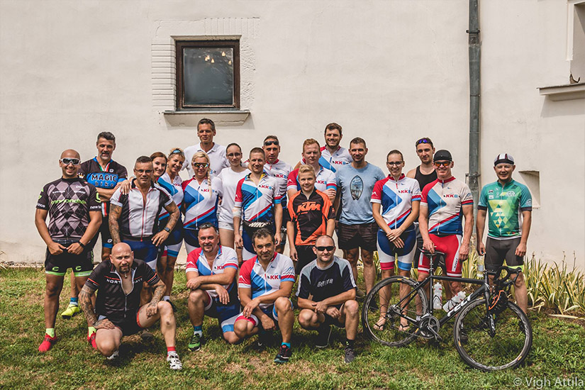 Magyarországi túra: Bringás csapat sportfelszerelésben pózol a szabadban fehér fal előtt, túrára kész kerékpárral a fűben. Tekeregj.hu