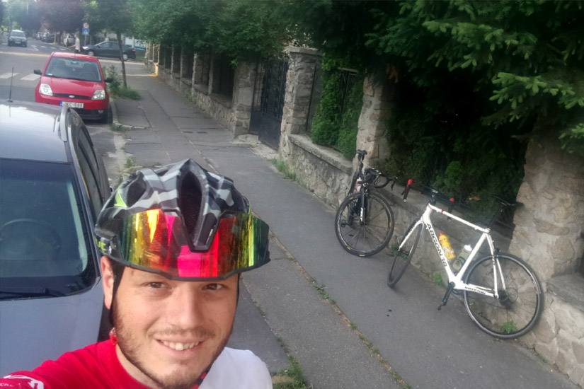 Magyarországi túra: Bringás férfi piros-fehér mezben selfie-t készít kiránduláson, mellette két kerékpár és fákkal szegélyezett utcai panoráma. Tekeregj.hu