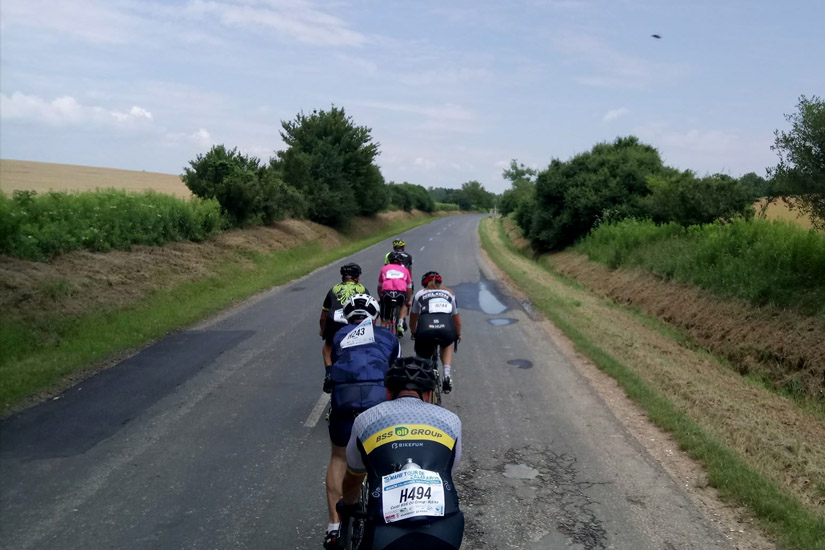 Magyarországi túra: Kerékpáros csoport teker vidéki úton, zöld mezők közt, panorámás kilátással, vezetőjük rózsaszín sisakot lát. Tekeregj.hu