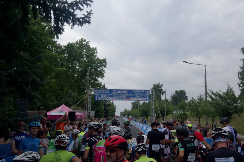 Magyarországi túra: Kerékpárosok rajtvonalnál, sisakban és számozott mezben, felhős ég alatt, fák és zászlók között. Tekeregj.hu